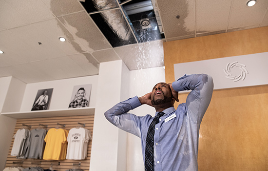A clothing store owner looks up disbelievingly at his heavily leaking ceiling with a hole in it, before commercial water damage restoration in Santa Ana
